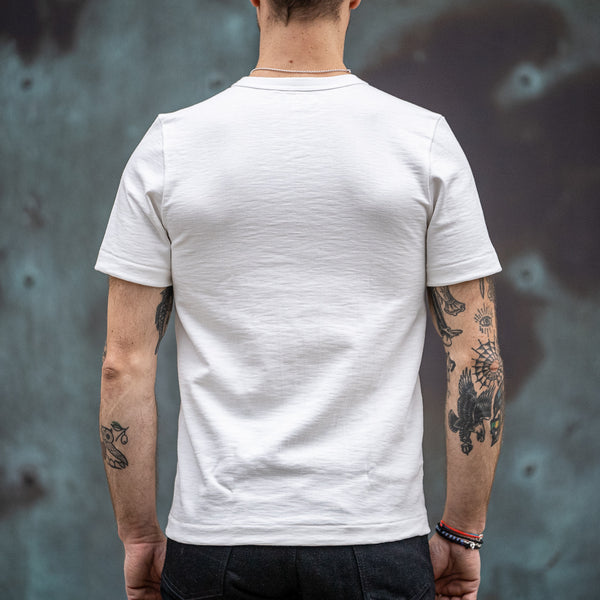 Ein Mann trägt ein weißes T-Shirt von Merz b. Schwanen und steht vor einer Wand. 
