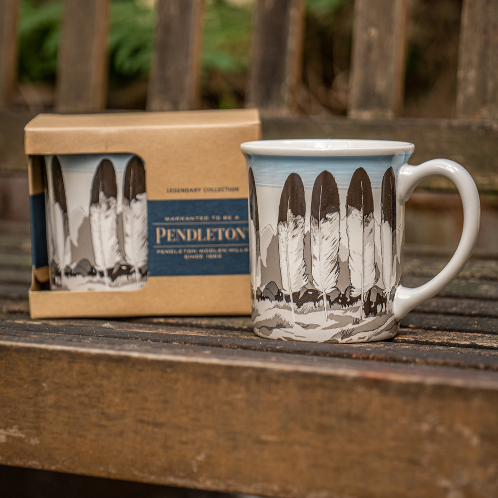 Zwei Keramiktassen von Pendleton mit Feder-Motiv auf einer Parkbank. 