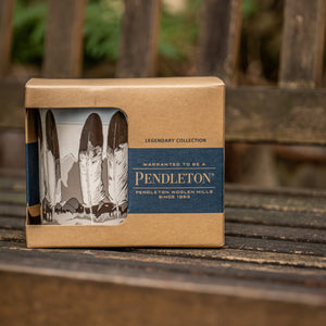 Eine Keramiktasse von Pendleton mit Feder-Motiv in einer Verpackung auf einer Parkbank. 